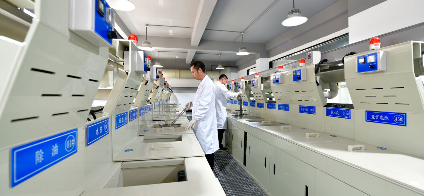 Two lab technicians in white coats work at a row of industrial equipment, possibly plating baths.