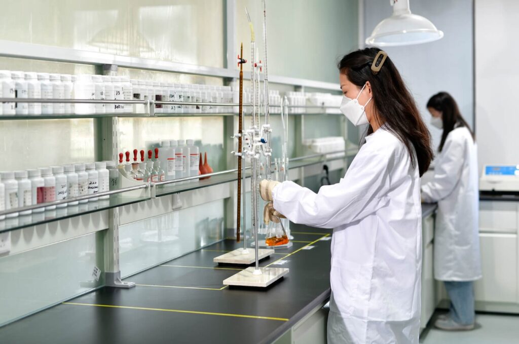 A scientist in a white lab coat and a face mask works in a laboratory, using glassware and equipment on a black countertop. Shelves filled with bottles and containers are visible in the background. Another scientist is visible in the background.