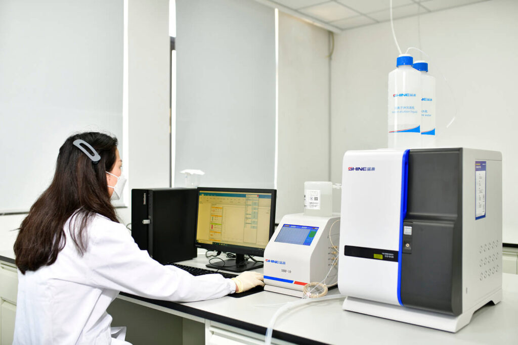 A scientist wearing a lab coat and face mask sits at a laboratory bench, working with a computer and specialized analytical equipment.