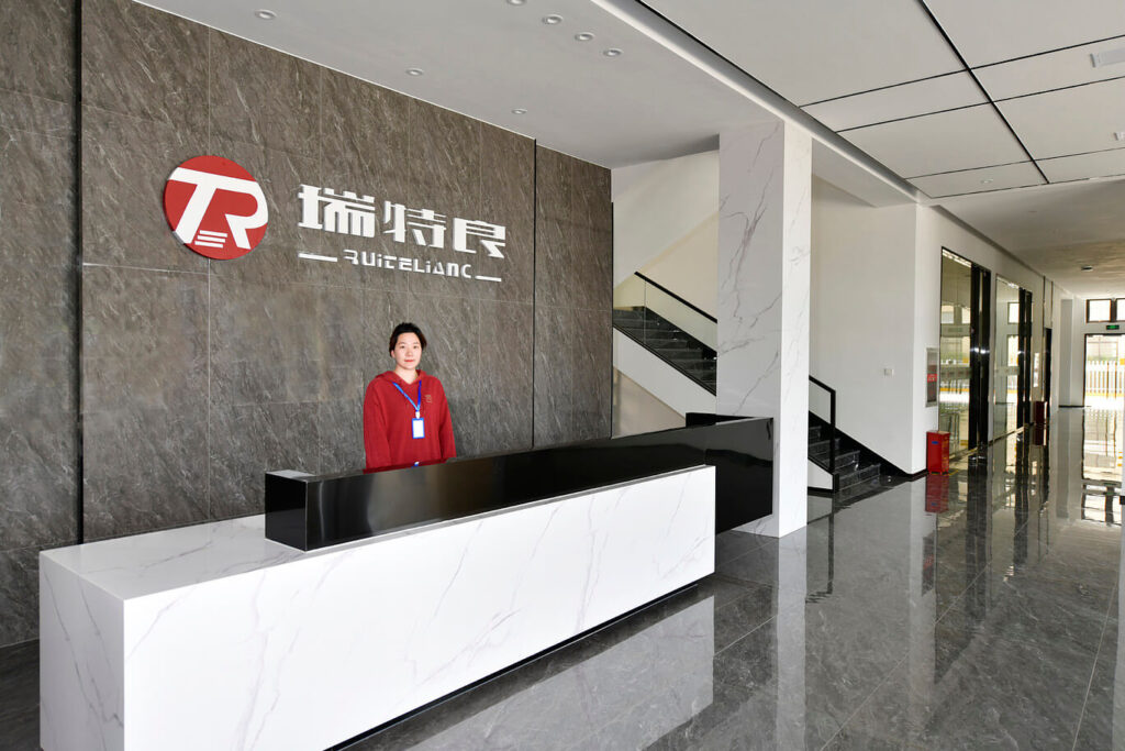 A modern office reception area with a white and black reception desk. A receptionist in a red and blue uniform stands behind the desk. The company logo, with a stylized "R" in a red circle, is displayed on the gray wall. A staircase is visible to the right.