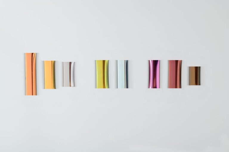 Small, rectangular pieces of metal in various metallic colors, including gold, silver, light blue, pink, and bronze, arranged in a horizontal row on a white background. They are likely samples of different finishes.