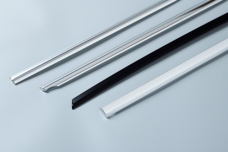 Four different colored and shaped trim pieces lie on a light gray background. Two of the trim pieces are silver, with one each of black, and white, and have varying profiles.