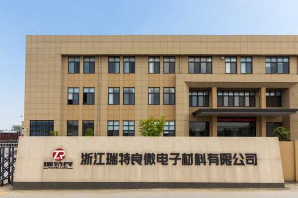 The exterior of a modern office building. The building facade is tan with multiple windows, and the company name and logo are displayed on a sign at the entrance.