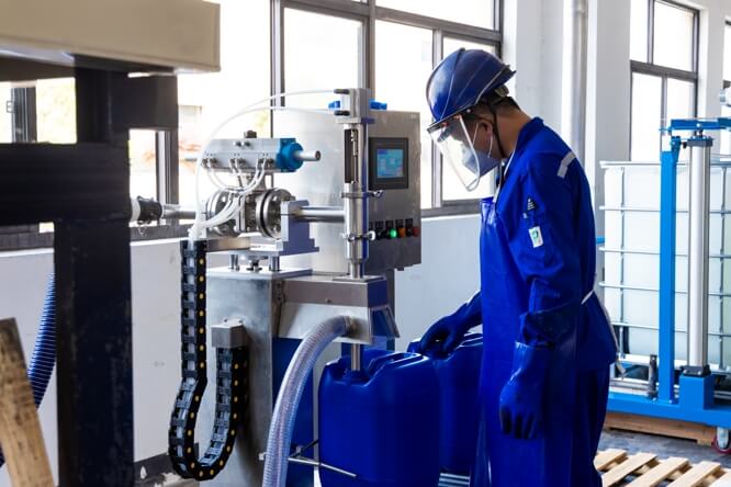 A worker in blue coveralls, gloves, a face mask, and a hard hat monitors a filling machine connected to blue plastic containers.