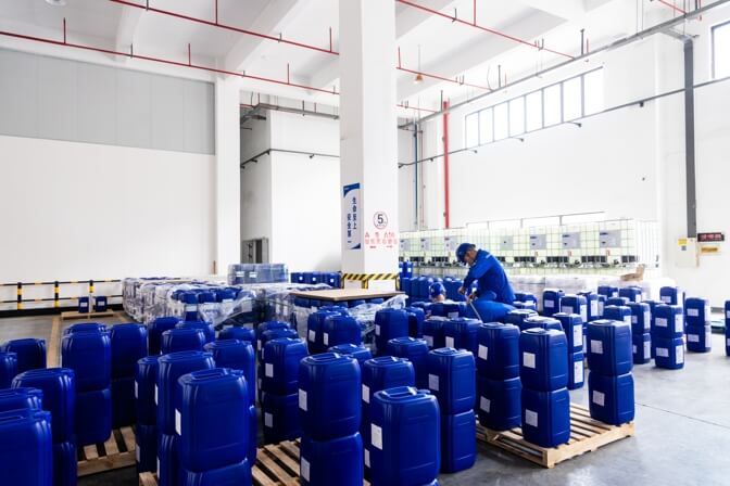 A factory worker in blue coveralls and a helmet uses a hand truck to move pallets of blue plastic containers in a warehouse.