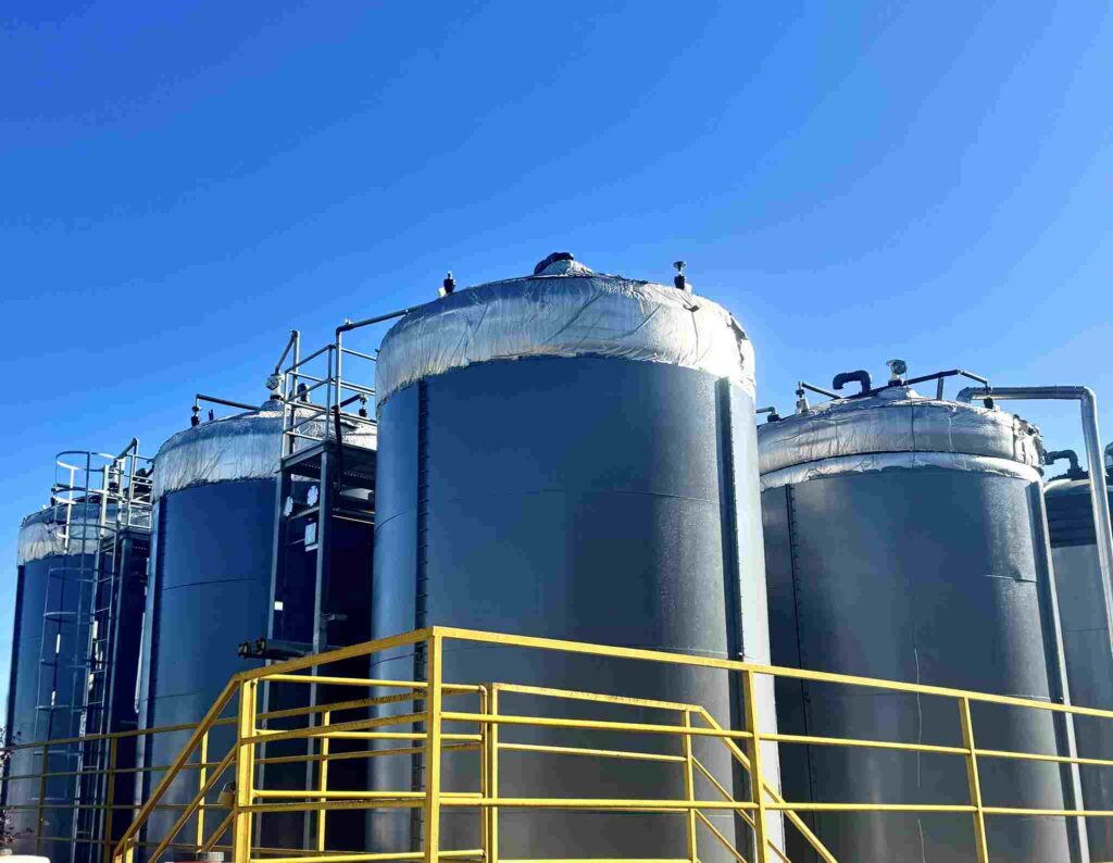 Three large industrial storage tanks under a clear blue sky. The tanks are gray, and their tops are covered with wrinkled, silver insulation. Yellow metal railings and ladders provide access to the tanks.