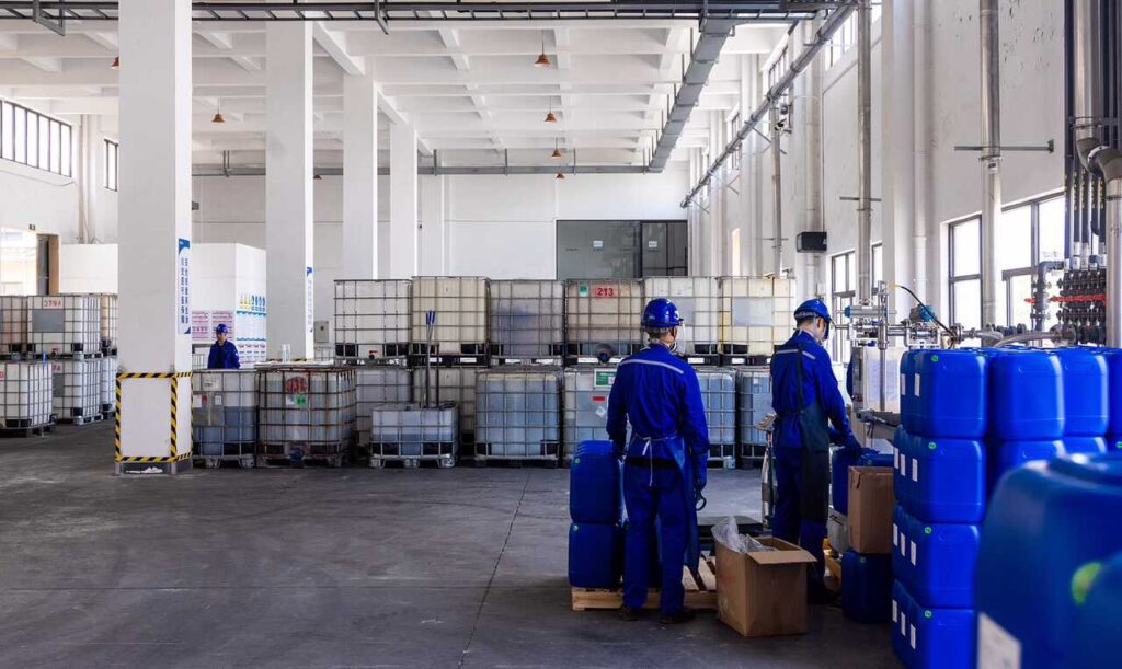 Workers in blue jumpsuits and hard hats are working in a warehouse with large white IBCs and smaller blue containers.