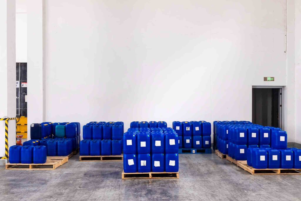 Stacks of blue plastic containers sit on pallets in a warehouse. A fire exit door is visible in the background.