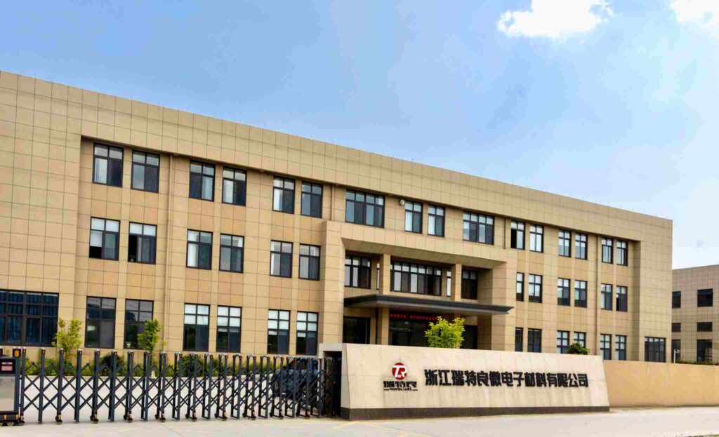 A modern, multi-story office building with tan siding and many windows. A retractable gate closes off the entrance, and a sign in front displays the RTL (Zhejiang Ruiteliang) logo.