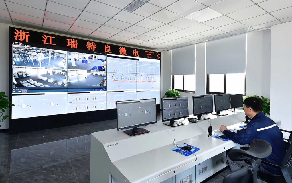 An industrial control room with a large video wall displaying various camera feeds and process data. A worker in a blue uniform sits at a desk with multiple computer monitors, reviewing information.