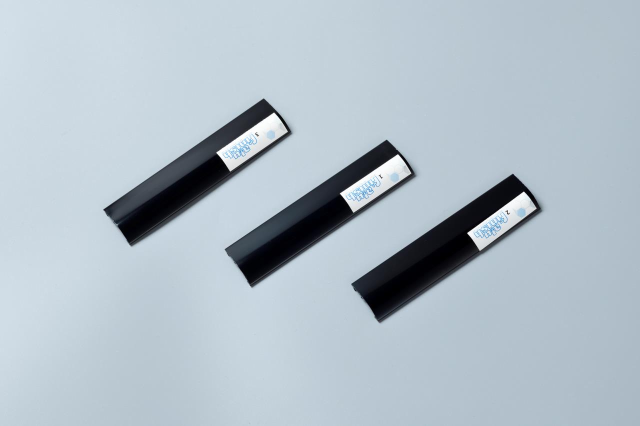 Three black plastic extrusion samples lie on a light gray background. Each sample has a small white label with illegible blue text.
