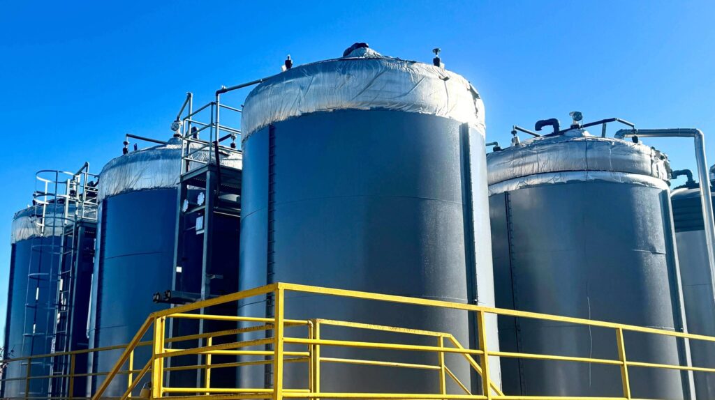 Three large gray industrial tanks against a vibrant blue sky. The tanks have silver insulation on their tops and are surrounded by yellow safety railings and ladders.