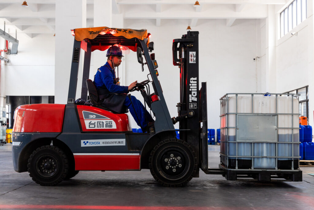 A worker in a blue jumpsuit and hardhat operates a red forklift carrying a large, gray IBC in a warehouse.
