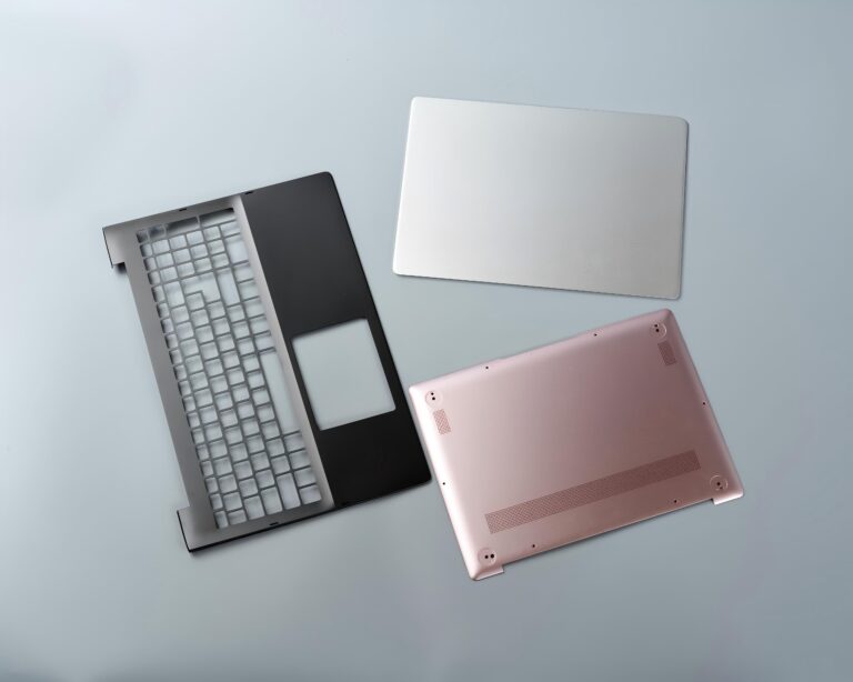 Laptop components, including a black keyboard deck, a rose gold bottom casing, and a silver trackpad cover, arranged on a light gray background.