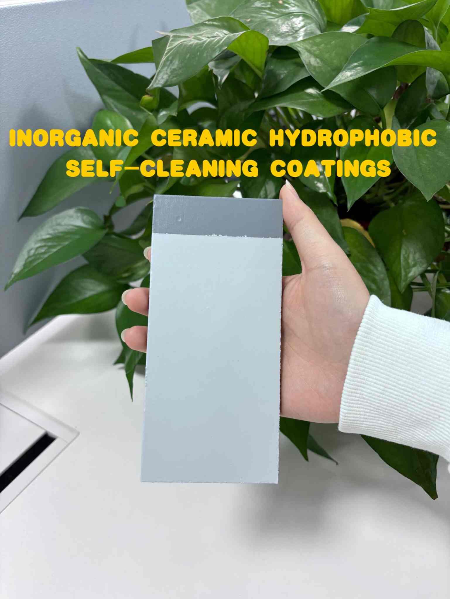 A hand holds a rectangular sample piece coated with inorganic ceramic hydrophobic self-cleaning coating. The coating is a light gray color, with a darker gray strip along the top edge.